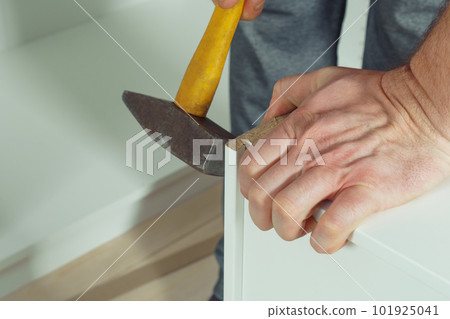 Male hands hold metal hammer with yellow plastic handle, driving dowel, assembling white drawer of dresser or wardrobe Male hands hold metal hammer with yellow plastic handle, driving dowel, assembling white drawer of dresser or wardrobe 101925041