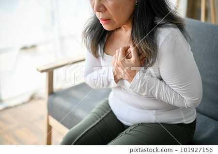 Unwell Asian mature woman sitting on sofa, suffering from heart attack or chest pain. 101927156