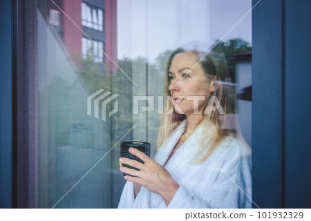 Woman in elegant robe drinking coffee in hotel room and standing near window. window reflection Woman in elegant robe drinking coffee in hotel room and standing near window. window reflection 101932329