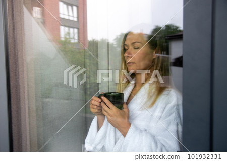 Woman in elegant robe drinking coffee in hotel room and standing near window. window reflection 101932331