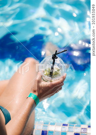 Female hand hold disposable cup with lemonade in swimming pool. Reflection of clear blue water. Summer mood Female hand hold disposable cup with lemonade in swimming pool. Reflection of clear blue water. Summer mood 101933849