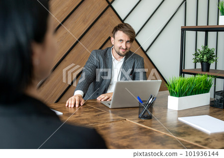 A cheerful man in a jacket is sitting at his desk in the office with a laptop. 101936144