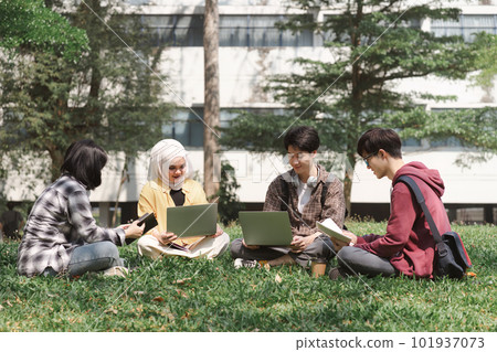 A multiethnic group of asian university students is seen studying together while sitting on the grass near the campus. 101937073