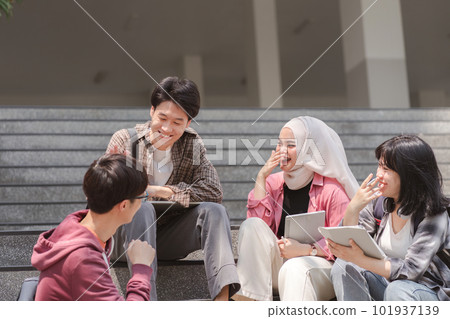 A group of diverse, multiracial young Asian university students are seen resting and chatting outdoors sitting on steps near the campus and communicating with each other. A group of diverse, multiracial young Asian university students are seen resting and chatting outdoors sitting on steps near the campus and communicating with each other. 101937139