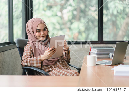 Beautiful muslim business woman wearing a hijab is diligently working at her tablet, displaying professionalism and commitment to her job. Beautiful muslim business woman wearing a hijab is diligently working at her tablet, displaying professionalism and commitment to her job. 101937247