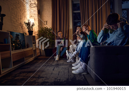 Group of friends, young people sitting on couch at home in the evening and singing karaoke, having fun, talking and drinking beer 101940015