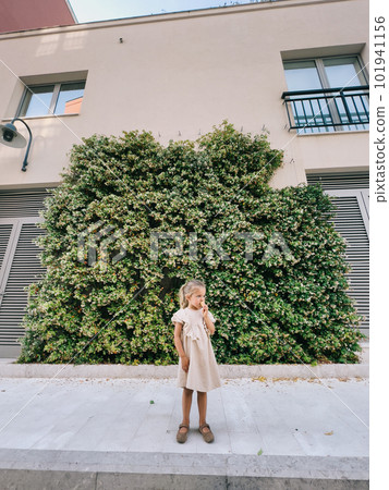 Little girl eating ice cream cone near the green hedge near the house 101941156