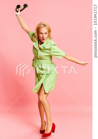 Portrait of charming happy woman, wearing green elegant costume holding headphones and dancing over pink studio background. Favourite song, music band 101941717