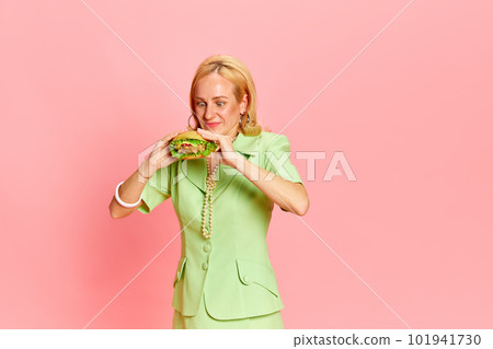 Portrait of beautiful blond woman holding in hands big fast food burger and looking on it with hunger over pink studio background. Lunch break 101941730