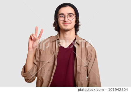 Positive curly man with little beard, makes peace sign, smiles pleasantly, dressed in fashionable hat and beige shirt, isolated over white background. Youth and body language concept. Boy says hello 101942804