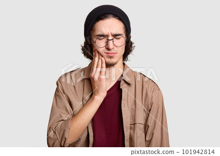Photo of stressful youngster with small beard keeps hand on cheek, suffers from toothache, keeps eyes shut, dressed in stylish clothes, big round spectacles, models against white studio wall. Photo of stressful youngster with small beard keeps hand on cheek, suffers from toothache, keeps eyes shut, dressed in stylish clothes, big round spectacles, models against white studio wall. 101942814