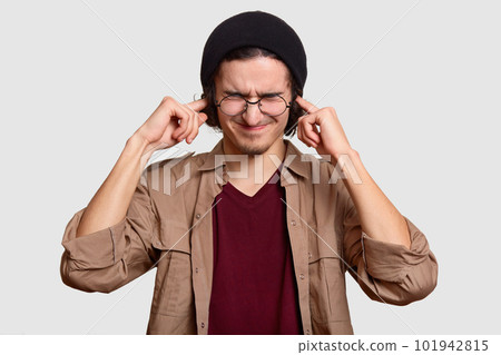 Stressful teenager wears black har and beige shirt, pluggs ears, ignores loud sound coming from noise neighbours, stands against white background. Frustrated hipster annoyed with something noisy Stressful teenager wears black har and beige shirt, pluggs ears, ignores loud sound coming from noise neighbours, stands against white background. Frustrated hipster annoyed with something noisy 101942815