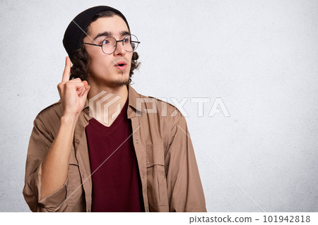 Image of young caucasian hipster man wearing glasses, maroon shirt, jacket and cap, posing isolated over white background, pointing index finger up with successful idea, copy space for advertisment. 101942818