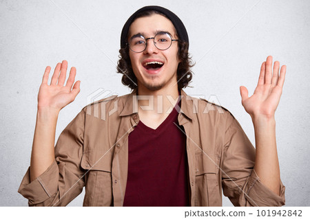 Cheerful attractive hipster standing isolated over white background in studio, raising his hands up, being in high spirits, wearing black hat, round spectacles, red sweatshirt, beige jacket. Cheerful attractive hipster standing isolated over white background in studio, raising his hands up, being in high spirits, wearing black hat, round spectacles, red sweatshirt, beige jacket. 101942842