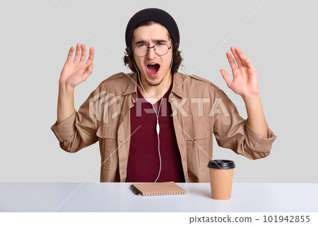 Depressed emotive guy raises hands and exclaims desperately, has annoyed facial expression, wears round optical spectacles, listens music, writes notes in notepad, isolated over white background Depressed emotive guy raises hands and exclaims desperately, has annoyed facial expression, wears round optical spectacles, listens music, writes notes in notepad, isolated over white background 101942855