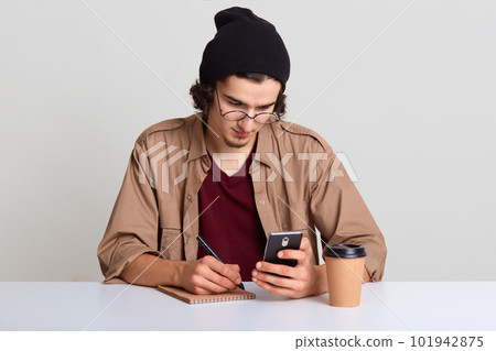 Studio shot of young man sitting at white desk and working, holding smart phone in hand and writting down something from it, looking concentrated on screen, drinks coffee. Student or freelance concept Studio shot of young man sitting at white desk and working, holding smart phone in hand and writting down something from it, looking concentrated on screen, drinks coffee. Student or freelance concept 101942875