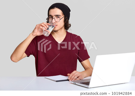 Handsome male freelancer feels thirsty, drinks water from glass, works on digital laptop computer, works from home, earns money online, wears casual t shirt and hat, isolated over white background 101942884