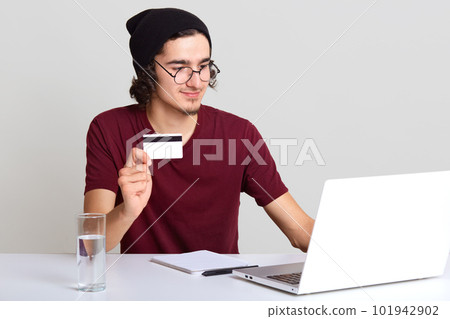Studio shot of male sitring at desk with opened laptop, showing blank card for your advertisement, uses electronic device for online payment, wearing maroon t shirt, black cap. Techology concept. 101942902