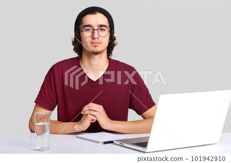 Relaxed man keeps hands together, looks at camera, tries to relax after work, sits at workplace, surrounded with portable laptop computer, notebook and glass of water, has curly hair isolated on white 101942910
