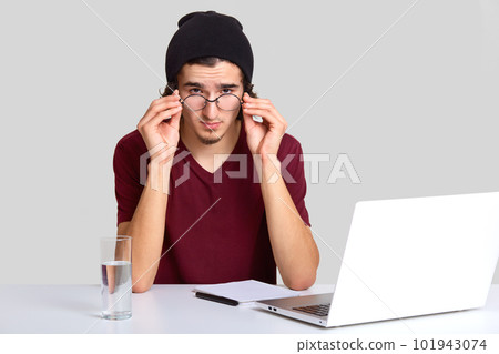 Confident student sits at workplace, looks attentively through round transparent glasses, dressed in stylish clothes, makes notes in notebook as listens webinar via laptop computer glass of water near 101943074