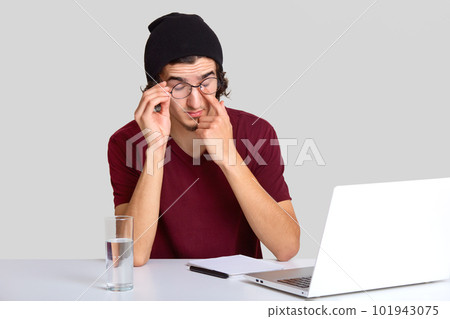 Photo of tired man rubbs eyes, wears glasses, has pain in eyes after long work at laptop computer, wears casual t shirt, prepares course work, uses modern portable laptop, isolated over white wall 101943075