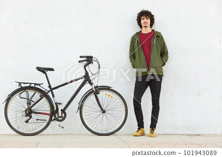 Pleased active young hipster being in riding tour, having stop to relax, standing near his bicycle, keeping his hands in pockets, being sport amateur, looking calm. Leisure time activities concept. 101943089