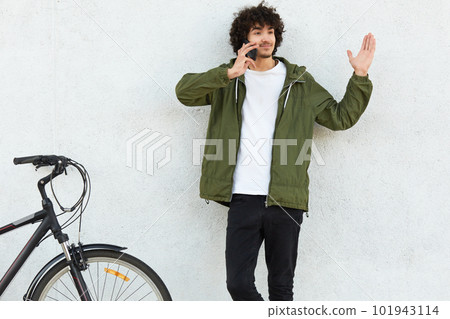 Puzzled hesitant guy has telephone conversation, raises hand with bewilderment, cant make choice while recieves suggestion, poses near bicycle against white background. So what to do, I dont know 101943114