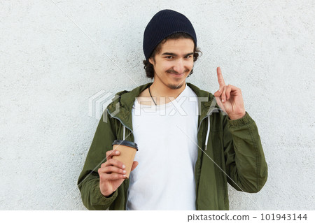 Portrait of young man with crisp hair and little beard, in casual anorak, black cap, holds takeaway coffee, pointing index finger up, isolated on white background, stands smiling, has interesting idea 101943144