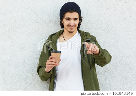 Handsome guy with pleased friendly expression, indicates directly at camera with index finger, dressed in fashionable jacket, warms himeseld during cold weather with hot beverage isolated on white 101943149