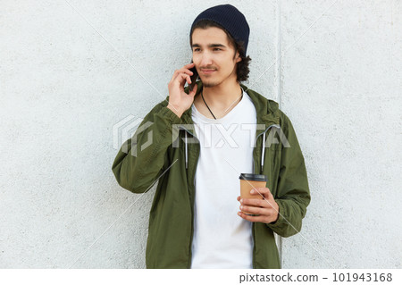 Slim pleasant boy looking aside, holding his smartphone near ear, concentrated on conversation, wearing white t shirt, green jacket and black hat, holding papercup of coffee. Youth and time concept. 101943168