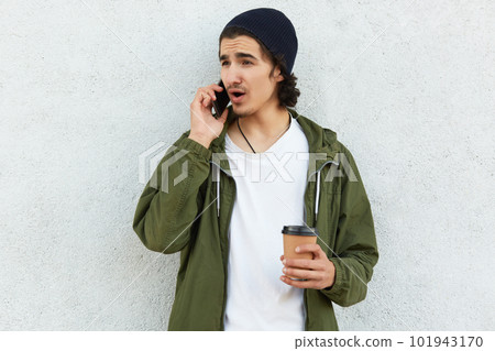 Indignant fashionable young guy has phone talk, uses modern electronic device, drinks takeaway coffee, focused aside, poses over white studio wall. People, coffee break and communication concept 101943170