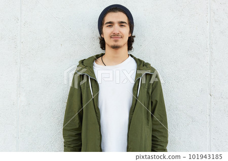 Horizontal shot of stylish teenage boy wears black hat, white t shirt and green anorak, looks with satisfied expression direclty at camera, isolated over white background. Lifestyle and people concept 101943185