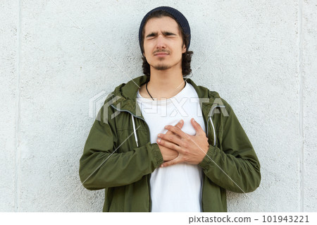 Portrait of suffering disappointed hipster putting his hands on heart, closing his eyes, heart attack, feeling pain, isolated over white wall outdoors alone. People and health problems concept. 101943221
