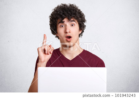 Portrait of surprised hipster guy keeps jaw dropped and fore finger raised, uses laptop computer, gets brilliant idea for creating new article, isolated over white concrete wall. People and technology Portrait of surprised hipster guy keeps jaw dropped and fore finger raised, uses laptop computer, gets brilliant idea for creating new article, isolated over white concrete wall. People and technology 101943243