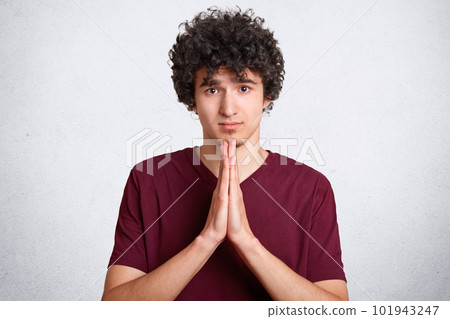 Please, forgive me! Handsome young curly male makes praying gesture, keeps palms pressed together, feels guilty, dressed in casual t shirt, asks for apologize, isolated over white background 101943247