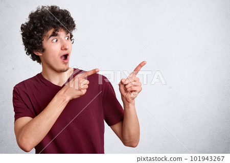 Portrait of impressed curly haired handsome guy raising hands, showing direction with forefingers, looking aside, wearing casual dark red t shirt, being shocked. Copy space for advertisement. Portrait of impressed curly haired handsome guy raising hands, showing direction with forefingers, looking aside, wearing casual dark red t shirt, being shocked. Copy space for advertisement. 101943267