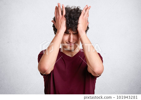 Attractive male with curly hair, wears casual maroon t shirt, has unhappy expresions, touches his head with hands isolated over white background. Copy space for your advertisment or promotional text. Attractive male with curly hair, wears casual maroon t shirt, has unhappy expresions, touches his head with hands isolated over white background. Copy space for your advertisment or promotional text. 101943281