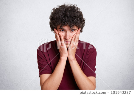 Close up portrait of serious man touches cheek with palm, looks directly at camera, has pensive expression, wears casual maroon t shirt, isolated over white background. People and emotions concept. Close up portrait of serious man touches cheek with palm, looks directly at camera, has pensive expression, wears casual maroon t shirt, isolated over white background. People and emotions concept. 101943287