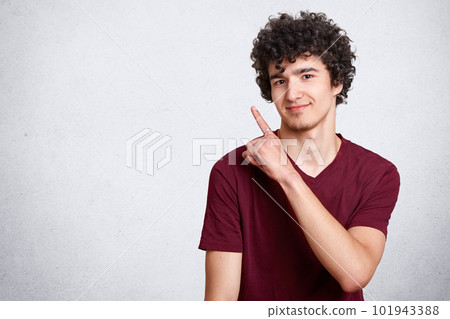 Portrait of stylish handsome young teenager isolated on white background. Dark haired curly student smiles and points aside. Attractive teen poses in photo studio. Copy space for your advertsment. Portrait of stylish handsome young teenager isolated on white background. Dark haired curly student smiles and points aside. Attractive teen poses in photo studio. Copy space for your advertsment. 101943388