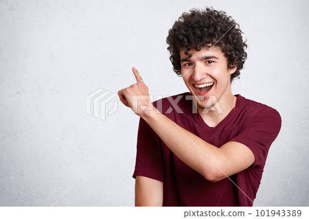 Portrait of stylish handsome young guy dresses casual outfit, with dark curly hair isolated on white background. Laughing teenager being in good mood points up. People, youth and lifestyle concept. Portrait of stylish handsome young guy dresses casual outfit, with dark curly hair isolated on white background. Laughing teenager being in good mood points up. People, youth and lifestyle concept. 101943389