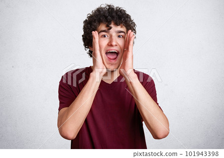 Emotional brunet male has dark curly hair, looks nervously directly at camera, finds someone, scream loudly, male wearing casual maroon t shirt, keeps hands near mouth, isolated over white background. Emotional brunet male has dark curly hair, looks nervously directly at camera, finds someone, scream loudly, male wearing casual maroon t shirt, keeps hands near mouth, isolated over white background. 101943398