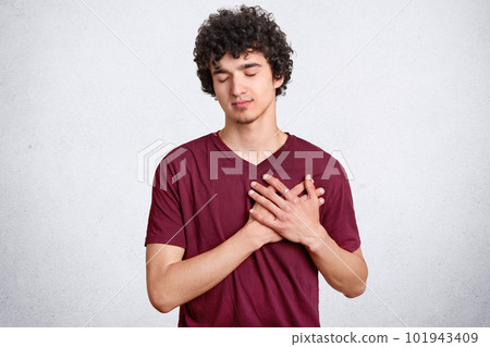 Thankful young boy keeps hands on heart, being touched by heart piercing story, keeps eyes closed, wears casual outfit, isolated over white concrete wall. People, youth and body language concept Thankful young boy keeps hands on heart, being touched by heart piercing story, keeps eyes closed, wears casual outfit, isolated over white concrete wall. People, youth and body language concept 101943409