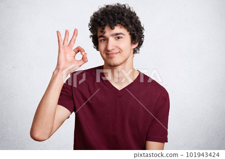 Young handsome male with curly hair, makes okay sign, shows his approval, gestures with hand, isolated over white background. Satisfied man demonstrates great sign, assures he will deal with work Young handsome male with curly hair, makes okay sign, shows his approval, gestures with hand, isolated over white background. Satisfied man demonstrates great sign, assures he will deal with work 101943424