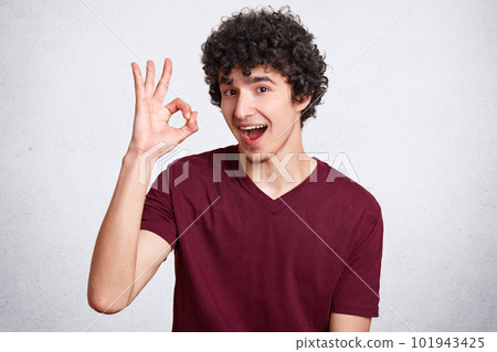 Everything will be fine! Satisfied curly man looks with assured and happy expression, shows ok sign, demonstrates his readiness and awareness, dressed in casual t shirt, isolated over white background Everything will be fine! Satisfied curly man looks with assured and happy expression, shows ok sign, demonstrates his readiness and awareness, dressed in casual t shirt, isolated over white background 101943425