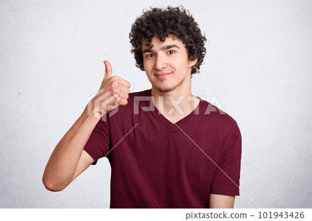 Handsome curly male with delighted expression, keeps thumb raised, shows his approval, poses against white concrete wall. Satisfied teenager with crisp hair shows ok sign. People and body language Handsome curly male with delighted expression, keeps thumb raised, shows his approval, poses against white concrete wall. Satisfied teenager with crisp hair shows ok sign. People and body language 101943426