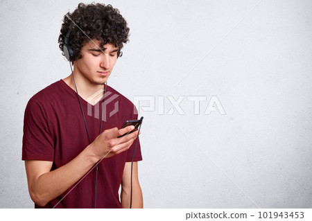 Horizontal shot of young handsome man, has dark curly hair, guy with cell phone in hand and earphones listening to music on white background with copy space for your advertismant and promotion text. 101943453