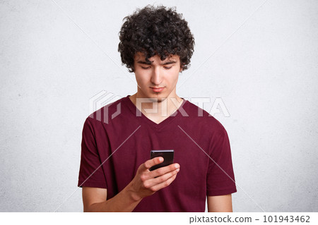 Good looking guy with concetrated look in mobile phone, recieves message with offer to go out, wears casual t shirt, poses against white concrete wall. Curly man uses modern technologies indoor Good looking guy with concetrated look in mobile phone, recieves message with offer to go out, wears casual t shirt, poses against white concrete wall. Curly man uses modern technologies indoor 101943462