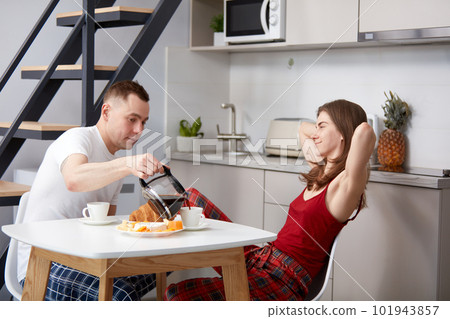 Happy young couple, man and woman having morning breakfast at home in kitchen. Husband pouring coffee to his smiling wife Happy young couple, man and woman having morning breakfast at home in kitchen. Husband pouring coffee to his smiling wife 101943857
