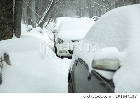 a row of parked cars on a city street on a cold winter day near the road among the trees, a snow blizzard covers vehicles. a row of parked cars on a city street on a cold winter day near the road among the trees, a snow blizzard covers vehicles. 101944198
