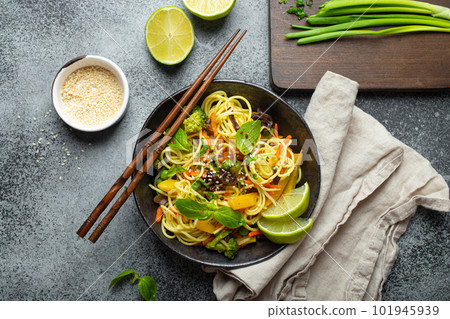 Asian vegetarian noodles with vegetables and lime in black rustic ceramic bowl, wooden chopsticks, cutting board with chopped green onion top view on stone background. Cooking noodles 101945939